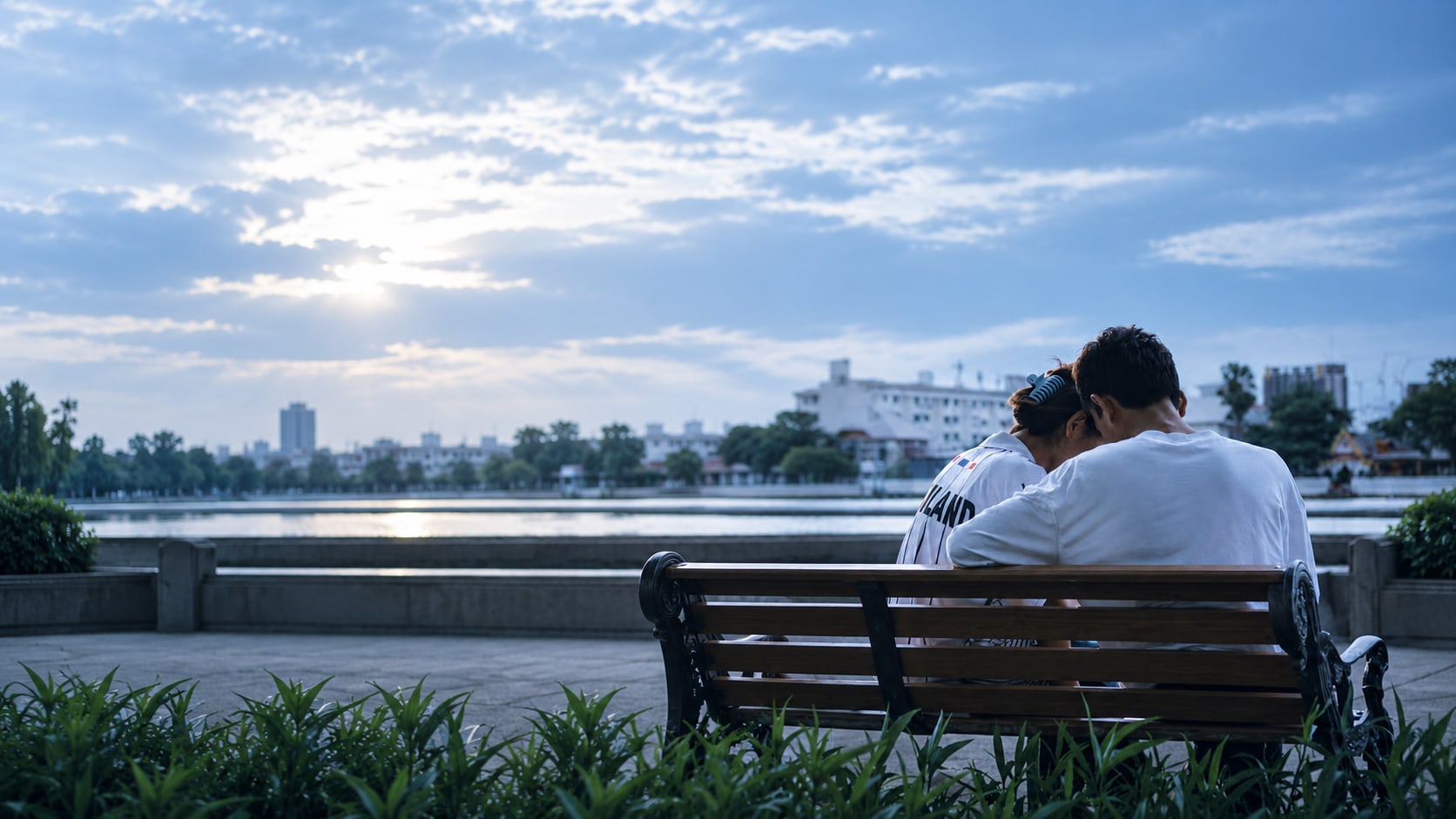 Two people sitting close together on a park bench at dusk, one leaning on the other's shoulder, illustrating the value of reaching out and being listened to